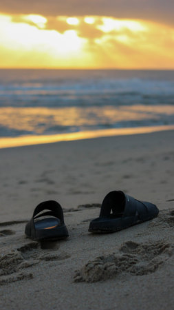 A pair of black sandals resting on a sandy beach during sunset. The golden sky reflects on the ocean waves as they gently wash ashore.のeditorial素材