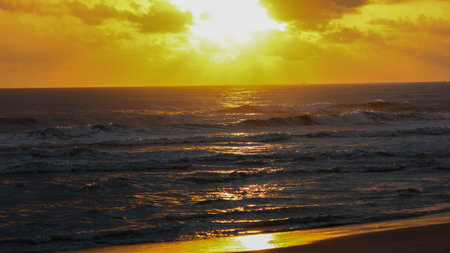 Golden light from the setting sun illuminating ocean waves and the wet sandy beach. Clouds are visible in the bright sky.のeditorial素材
