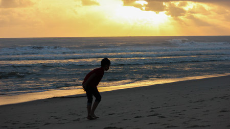 A young child stands silhouetted on a sandy beach during sunset, with the golden sun reflecting on the ocean waves and a cloudy sky overhead.のeditorial素材
