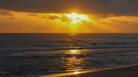 A vibrant golden sunrise over the ocean, with rays of sun breaking through dark clouds. The sun's reflection creates a shimmering path on the water, leading to a dark sandy beach.のeditorial素材