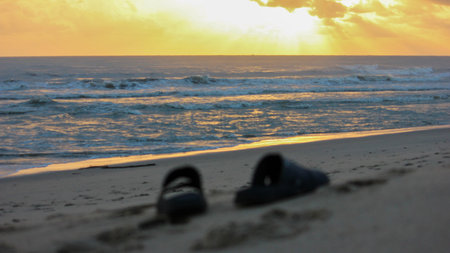 A pair of dark slippers left on a sandy beach during sunset, with waves crashing on the shore in the background. The sky is filled with warm orange and yellow hues from the setting sun.のeditorial素材