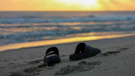 A pair of black sandals sits on a sandy beach during sunset, with gentle ocean waves and a vibrant orange and yellow sky forming the background.のeditorial素材