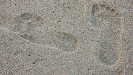 Close-up view of two distinct human footprints pressed into the wet sand of a beach. The sand is light beige with small grains.のeditorial素材