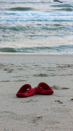A pair of bright red children's clogs are left on the wet sand of a beach, with gentle ocean waves visible in the blurry background.のeditorial素材