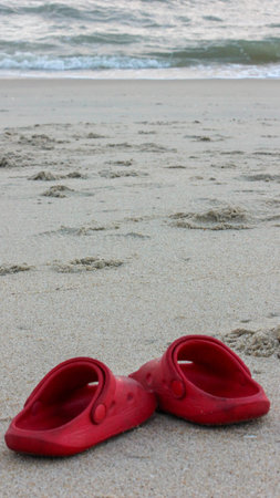 A pair of vibrant red clogs rests on the light-colored sand of a tranquil beach, with soft ocean waves breaking gently in the blurred background. The image suggests a peaceful moment of relaxation or a vacation by the sea.のeditorial素材