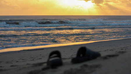 Golden sunrise over the South China Sea with waves breaking on a sandy beach. Two dark sandals are visible in the foreground, partially out of focus.のeditorial素材