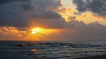 A vibrant sunrise over the ocean, with the sun breaking through dark, heavy clouds, casting bright orange and yellow light across the sky and reflecting on the water's surface and gentle waves approaching a sandy beach.のeditorial素材