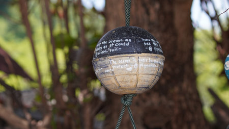 A close-up view of a spherical buoy, painted black and beige, with handwritten messages in white chalk promoting ocean conservation and protection. The buoy is suspended by a green rope amidst a blurred background of green foliage and brown tree trunks. Messages include 'Keep the ocean as bounty as it could' and 'The ocean is ours to share and protect'.の写真素材