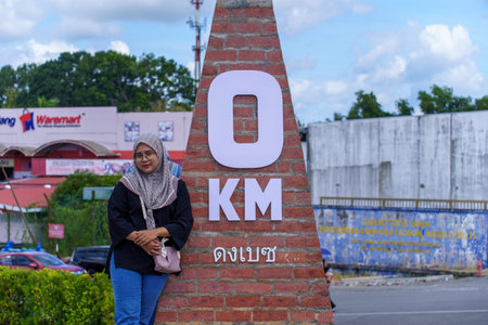 A young woman wearing a hijab and glasses poses next to a brick '0 KM' distance marker, with the 'Waremart' store visible in the background under a cloudy sky in Kangar, Perlis, Malaysia.のeditorial素材