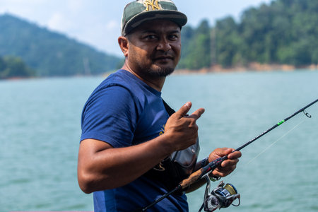 A Malaysian man, wearing a blue t-shirt and a New York Yankees baseball cap, is captured outdoors while fishing. He is holding a fishing rod with a spinning reel, looking directly at the camera, and making a pointing gesture. The background shows tranquilのeditorial素材
