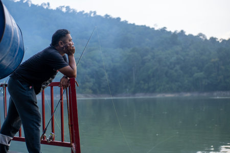 A man takes a moment to relax and fish by a large freshwater lake surrounded by lush green mountains in Malaysia. He leans on a red railing, holding a fishing rod, and gazes out at the calm water and misty forest landscape, enjoying the peaceful outdoor aのeditorial素材