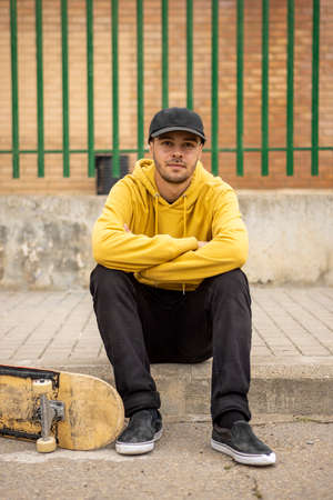 Young male in a yellow hoodie and cap with a skateboard sitting on the groundの写真素材