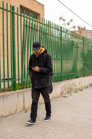 Young male with a black coat and cap walking on the street looking at the phoneの写真素材