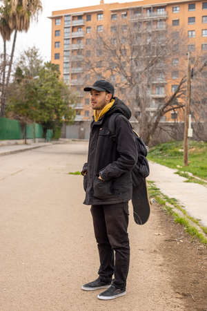 Young male with a black coat and cap standing on the street with a skateboardの写真素材