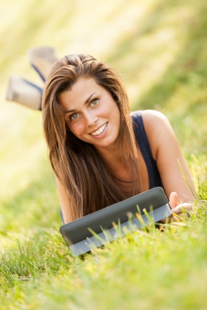Pretty brunette girl is looking a digital screen while sitting on the grass and smilingの写真素材