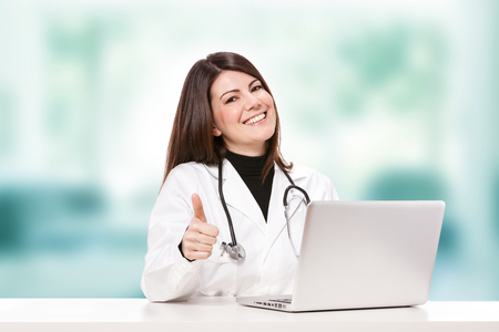 Image of female doctor sitting at her desk and smiles towards the cameraの写真素材