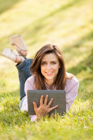 pretty brunette woman laying on a grass field while looking on a digital tabletの写真素材
