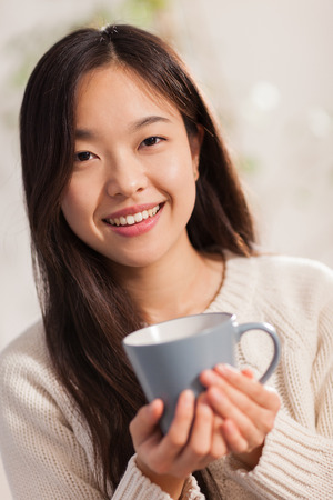 young asian woman with hot mug in her hands smiling towards the cameraの写真素材