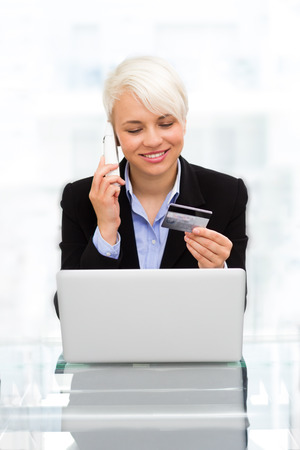 photo blonde smiling woman speaking at phone while online shopping with credit card in officeの写真素材