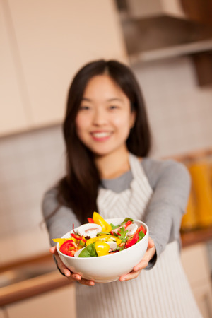 photo of asian smiling woman holding a colorful salad in her hands in the kitchenの写真素材