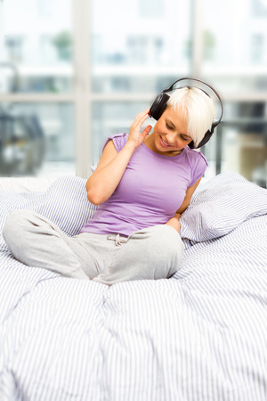 Blonde smiling woman is listening to music with headphones in the bedの写真素材