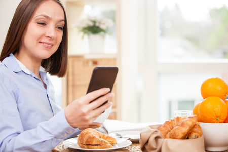 Smiling brunette businesswoman is having breakfast while checking her emails at homeの写真素材