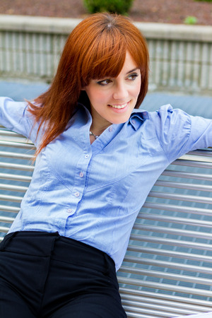 Photo of businesswoman with red hair sitting outside on a metal bench while smilingの写真素材