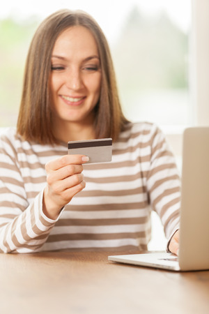 Young smiling woman holding a credit card in her hand while doing online shopping at homeの写真素材