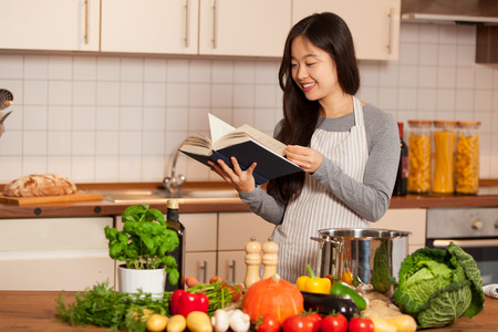 Asian smiling woman looking a cookbook while standing in her kitchen