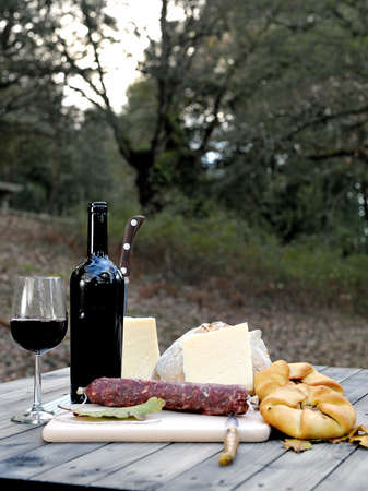 Bread, cheese, sausage, red wine, glass, cutting board and knife arranged on a wooden table for a snack in the countryside.の写真素材