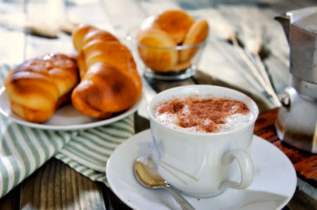 Close-up of a cup of coffee with milk, croissants, cookies and the coffee pot on an antique wooden table.の写真素材