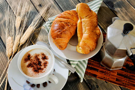 Close-up of a cup of coffee with milk, croissants and the coffee pot on an antique wooden table.の写真素材
