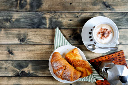 Top view of a cup of coffee with milk, croissant, brioche and coffee pot on an antique wooden table.の写真素材