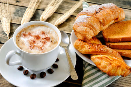 Close-up of a cup of coffee with milk, croissant, brioche and rusks on an antique wooden table.の写真素材