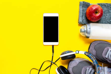 Top view of a red apple, sport shoes, audio headphone, smartphone, towel and water bottle photographed on yellow background. Text space.の写真素材