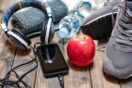 Close-up of a red apple, sport shoes, audio headphone, smartphone, towel and water bottle photographed on an antique wooden table. の写真素材