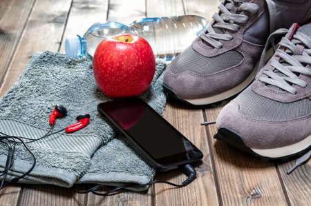 Close-up of a red apple, sport shoes, audio headphone, smartphone, towel and water bottle photographed on an antique wooden table. の写真素材
