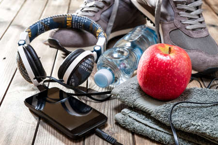 Close-up of a red apple, sport shoes, audio headphone, smartphone, towel and water bottle photographed on an antique wooden table. の写真素材