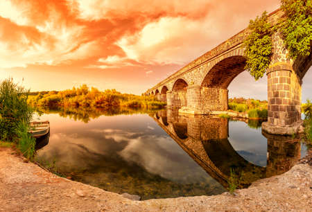 Beautiful sunset landscape of the Cedrino river with an old fishing boat and view on the Old Bridge of Oroseiの写真素材