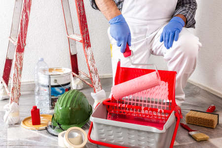 Caucasian house painter worker in white work overalls, dip the roller into the container with the paint. Construction industry.の写真素材
