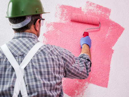 Caucasian house painter worker in white work overalls, with the roller he paints the wall with the colored painting of pink. Construction industry. Work safety.の写真素材