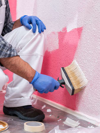 Caucasian house painter worker in white overalls, with the brush painting the pink wall with white paint. Construction industry. Work safety.の写真素材