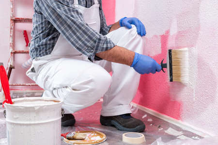 Caucasian house painter worker in white overalls, with the brush painting the pink wall with white paint. Construction industry. Work safety.の写真素材