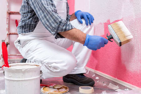 Caucasian house painter worker in white overalls, with the brush painting the pink wall with white paint. Construction industry. Work safety.の写真素材
