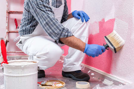 Caucasian house painter worker in white overalls, with the brush painting the pink wall with white paint. Construction industry. Work safety.の写真素材