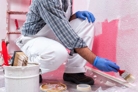Caucasian house painter worker in white overalls, painting the pink wall with white paint using a small brush. Construction industry. Work safety.の写真素材
