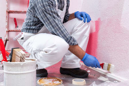 Caucasian house painter worker in white overalls, painting the pink wall with white paint using a small brush. Construction industry. Work safety.の写真素材
