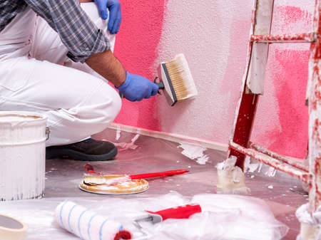 Caucasian house painter worker in white overalls, with the brush painting the pink wall with white paint. Construction industry. Work safety.の写真素材