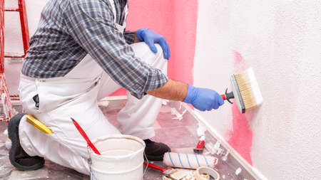Caucasian house painter worker in white overalls, with the brush painting the pink wall with white paint. Construction industry. Work safety.の写真素材