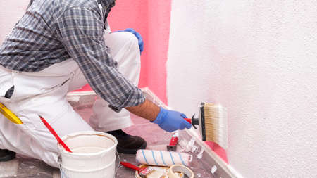 Caucasian house painter worker in white overalls, with the brush painting the pink wall with white paint. Construction industry. Work safety.の写真素材
